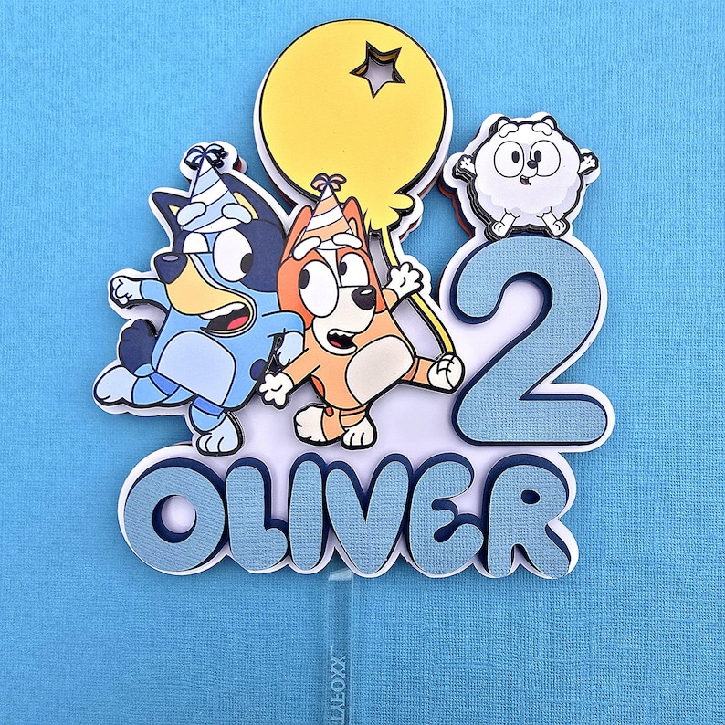 May include: Blue, orange, and white cartoon characters on a blue and white cake topper with the number 2 and the name Oliver.