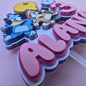 May include: A pink, blue, and white cake topper with the name "Alana" and a number "3" on it. The topper features cartoon characters from the show "Bluey".