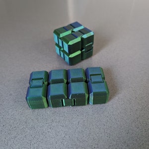 May include: Two green and blue 3D printed fidget toys. One is a cube and the other is a long rectangular shape.
