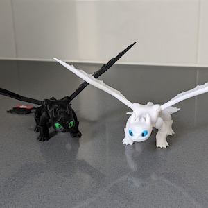 May include: Two toy dragons, one black with green eyes and one white with blue eyes. Both dragons have wings and are posed in a flying position.