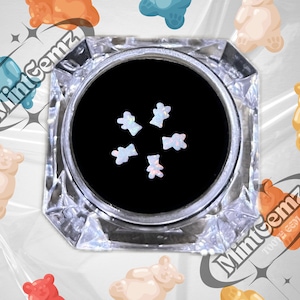 May include: A small clear container with a black lid filled with iridescent white bear-shaped nail charms. The container is surrounded by gummy bear-shaped stickers with the text "MintCenz Too Fly Gemz".