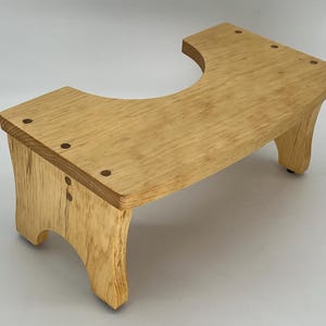 May include: A wooden toilet seat riser with a curved cutout and a natural wood finish. The riser has four legs and is designed to help people reach the toilet more easily.