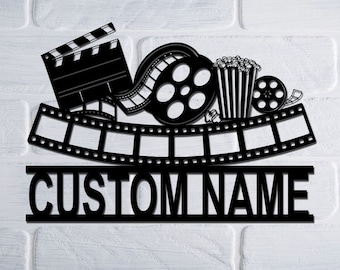 Pode incluir: Decoração de parede de metal preto com um rolo de filme, um balde de pipoca e uma claquete. O texto "CUSTOM NAME" é exibido abaixo do rolo de filme.