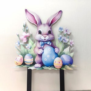 May include: A pastel pink and purple bunny rabbit holding a blue Easter egg with white polka dots. The bunny is surrounded by green leaves and pink and purple flowers. There are three additional Easter eggs in the scene, one pink and white, one blue and white, and one yellow and orange.