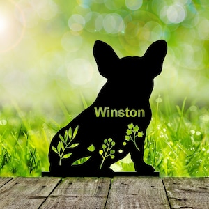 May include: Black silhouette of a French Bulldog with the name "Winston" written on its chest. The silhouette is set against a green grassy background with a wooden surface in the foreground.