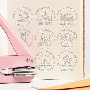 May include: A pink book embosser with a silver base is positioned next to a book page with several circular embossed library stamps. The stamps feature floral and book designs, along with names like Katie Roberts and Timothy Powell.