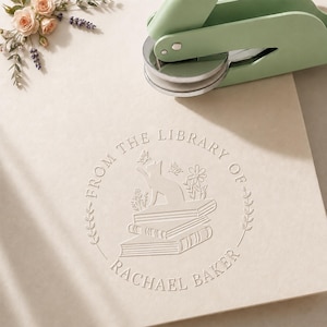 Peut inclure: Un outil de gaufrage en métal vert clair posé sur du papier crème. Le papier présente un motif gaufré circulaire avec le texte "FROM THE LIBRARY OF RACHEL BAKER" entourant une illustration d'un chat assis sur une pile de livres.