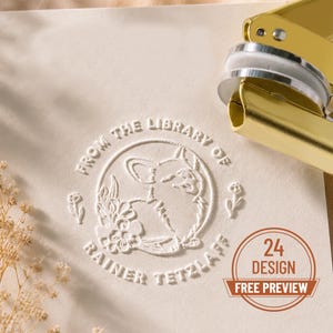 May include: A gold metal embosser tool positioned next to a cream-coloured paper featuring an embossed circular design. The design includes a corgi, floral elements, and the text "FROM THE LIBRARY OF RAINER TETZLAFF". A "24 DESIGN FREE PREVIEW" badge is visible.