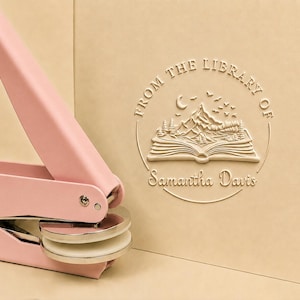 Peut inclure: Un outil de gaufrage en métal rose clair placé à côté d'une surface en papier beige avec un motif circulaire en relief. Le design comprend un livre ouvert avec des montagnes, des arbres, un croissant de lune, des oiseaux et le texte FROM THE LIBRARY OF Samantha Davis.