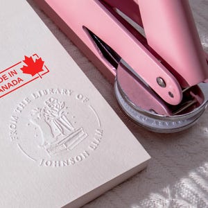 May include: A pink embosser rests on a white paper with a stamped design and the text "FROM THE LIBRARY OF JOHNSON LIAM." A red stamp reads "MADE IN CANADA" with a maple leaf. The embosser has a silver base.