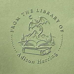 Peut inclure: Un motif de tampon gaufré vert sauge représentant un livre ouvert, une chaîne de montagnes et un dragon en vol. Le texte circulaire indique FROM THE LIBRARY OF Adison Herring. Le design est rendu dans un style en relief monochrome.