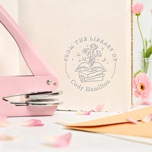 Peut inclure: Un gaufrage de livre rose avec une matrice argentée est à côté d'une page de livre crème avec un motif circulaire qui dit "From the Library of Cody Hamilton". Le motif représente un livre ouvert avec des fleurs. Des pétales de fleurs roses et une fleur rose sont en arrière-plan.
