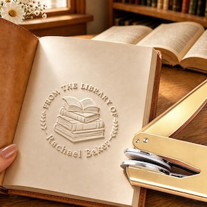 Peut inclure: Un outil de gaufrage en métal doré posé sur un livre ouvert à couverture en cuir beige. La page présente un motif circulaire en relief représentant une pile de livres avec le texte FROM THE LIBRARY OF Rachael Baker en bordure.