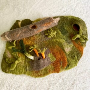 Könnte beinhalten: Eine Spielmatte aus Filz mit einem Landschaftsdesign in Grün-, Braun- und Orangetönen. Die Matte hat einen Baumstamm, Karotten und andere Details aus Filz. Die Matte ist für fantasievolles Spielen konzipiert.