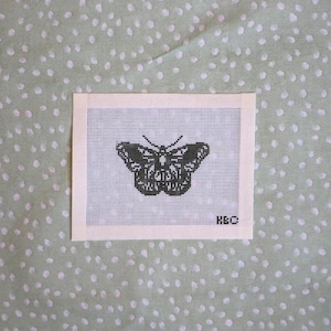 May include: A needlepoint canvas featuring a black butterfly design. The butterfly is centered on a white grid, with the initials "H&C" in the lower right corner. The canvas is set against a light green dotted background.