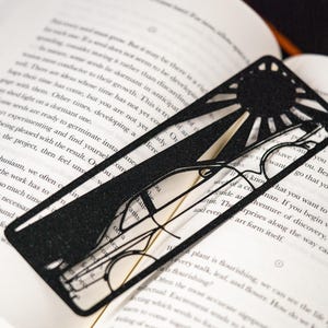 May include: A black, rectangular metal bookmark featuring a laser-cut design of a sun positioned above a pair of eyeglasses. The bookmark has a textured, glittery finish and is placed flat against the open pages of a white paper book.