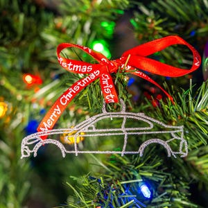 May include: A silver car-shaped Christmas ornament hanging on a decorated tree. The ornament is outlined with glitter and has a red ribbon with "Merry Christmas" printed on it. The tree is adorned with colorful lights.