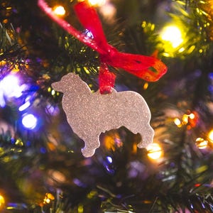 Puede incluir: Un adorno de perro de purpurina plateada colgado de un árbol de Navidad con una cinta roja y luces parpadeantes.