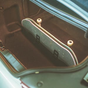 May include: A skateboard with a grey grip tape surface and white wheels positioned upright inside the dark carpeted trunk of a vehicle. The skateboard is secured in place by two black mounting brackets attached to the interior lining of the trunk.