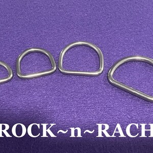 May include: Four silver metal D-rings of different sizes. The rings are arranged in a row from smallest to largest. The background is a purple fabric.