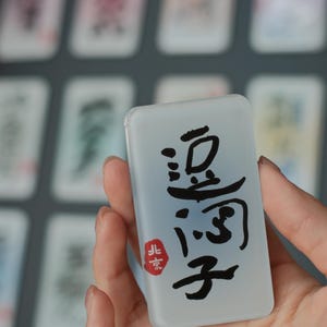 May include: A rectangular, translucent tile with black Chinese characters and a red stamp. The tile is held in a hand, with other tiles visible in the background. The characters are in a bold, calligraphic style.