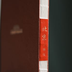 May include: A bound book with a reddish-brown cover and white stitching along the spine. A red strip on the spine features gold text. The book's pages are visible, and the overall aesthetic is classic and elegant.
