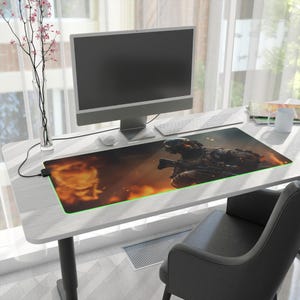 Peut inclure: Tapis de souris rectangulaire pour le jeu, représentant un soldat armé sur un fond enflammé. Le tapis a un bord éclairé en vert et est posé sur un bureau blanc avec un ordinateur, un clavier et une souris.