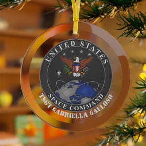Op de afbeelding: Een glazen kerstbal met een gouden rand en een zwart-wit afbeelding van het logo van het United States Space Command. De tekst "TSGT GABRIELLA GALLOSO" is onder het logo gedrukt.
