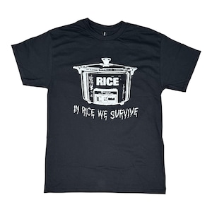 May include: A black short-sleeve t-shirt featuring a white graphic of a rice cooker with the word RICE printed on the front. Below the appliance, the text IN RICE WE SURVIVE is printed in a distressed white font across the chest area.