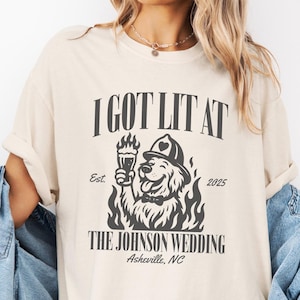 May include: Ivory-colored t-shirt with a graphic design. The design features a dog wearing a firefighter's hat, holding a beer, and surrounded by flames. Text on the shirt reads "I GOT LIT AT THE JOHNSON WEDDING Asheville, NC" and "Est. 2025".