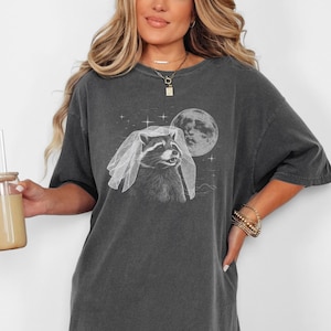 May include: Dark gray t-shirt featuring a raccoon wearing a veil, with a full moon and stars in the background. The shirt has a relaxed fit and is made of a soft material. The word "PEPPER" is printed at the bottom of the image.