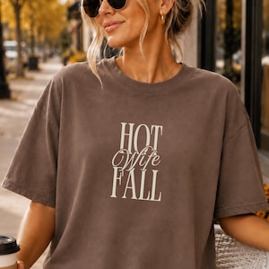 May include: A brown short-sleeved t-shirt featuring the text HOT Wife FALL printed in a cream-colored serif and script font on the center of the chest. The garment has a crew neckline and a relaxed fit with dropped shoulder seams.
