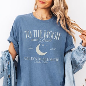 May include: A blue jean t-shirt with the text "To the Moon and Back" and a crescent moon graphic. The text also includes "June 2024" and "Ashley's Bachelorette Dallas, Texas".