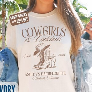 May include: Ivory-colored t-shirt with the text "COWGIRLS & Cocktails" in brown script. The design includes a cowboy hat, boots, and cocktail glasses. The text "ASHLEY'S BACHELORETTE Nashville, Tennessee" is also printed on the shirt.