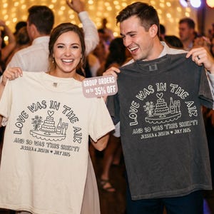 May include: A cream-colored t-shirt and a charcoal gray t-shirt, each featuring a white graphic of a wedding cake, flowers, and champagne flutes. The text reads: LOVE WAS IN THE AIR AND SO WAS THIS SHIRT! JESSICA & JUSTIN JULY 2025.