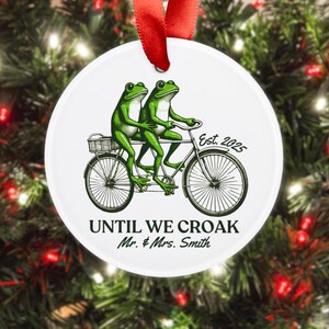 May include: White ceramic ornament with a red ribbon. The ornament features two green frogs riding a bicycle. Text includes "Est. 2025" and "UNTIL WE CROAK Mr. & Mrs. Smith". The ornament is hanging in front of a Christmas tree.