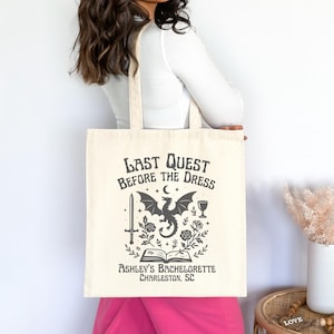 K&ouml;nnte beinhalten: Cremefarbene Tragetasche mit dem Text "Last Quest Before The Dress" und einer schwarzen Grafik eines Drachen, Schwerts und anderer Elemente. Der Text "Ashley's Bachelorette Charleston, SC" ist ebenfalls auf der Tasche aufgedruckt.