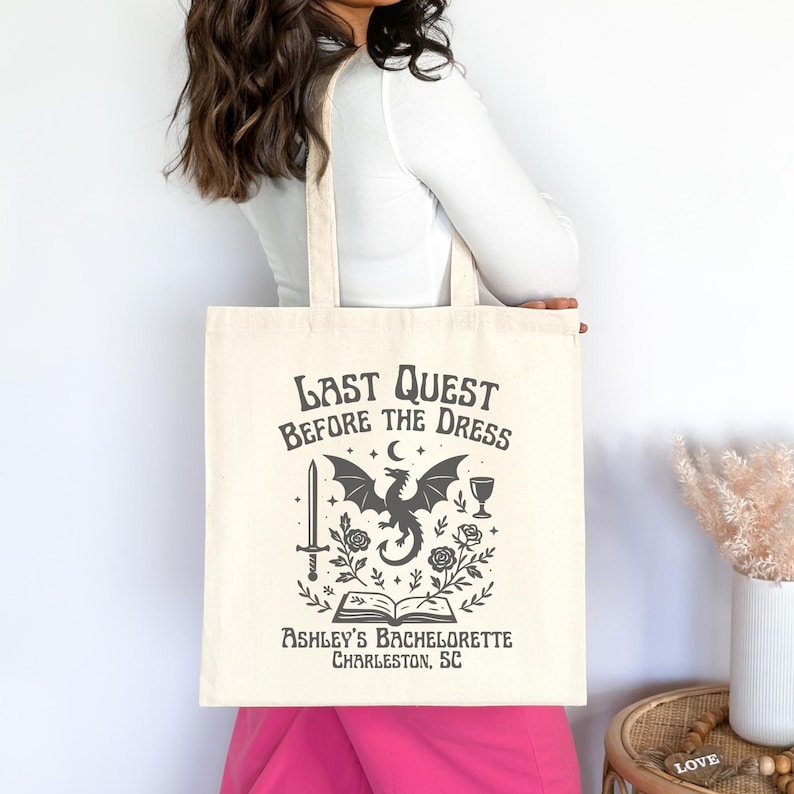 K&ouml;nnte beinhalten: Cremefarbene Tragetasche mit dem Text "Last Quest Before The Dress" und einer schwarzen Grafik eines Drachen, Schwerts und anderer Elemente. Der Text "Ashley's Bachelorette Charleston, SC" ist ebenfalls auf der Tasche aufgedruckt.