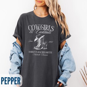 May include: Dark gray t-shirt with the text "COWGIRLS & Cocktails" and "ASHLEY'S BACHELORETTE Nashville, Tennessee". The design includes a cowboy hat, boots, and a cocktail glass. The shirt is a relaxed fit.