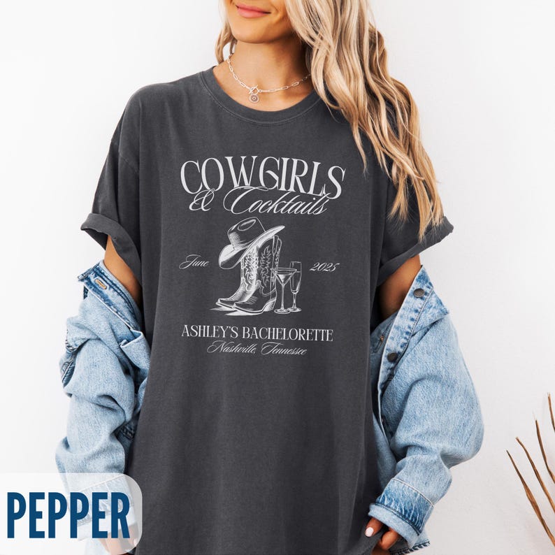 May include: Dark gray t-shirt with the text "COWGIRLS & Cocktails" and "ASHLEY'S BACHELORETTE Nashville, Tennessee". The design includes a cowboy hat, boots, and a cocktail glass. The shirt is a relaxed fit.