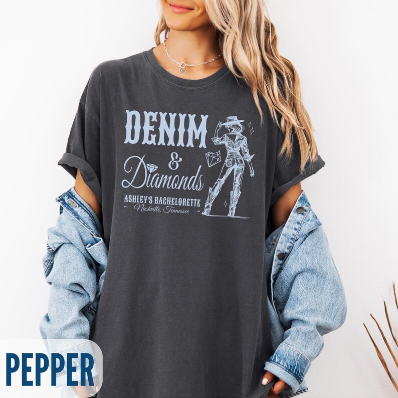 Puede incluir: Una camiseta gris oscuro con un dise&ntilde;o gr&aacute;fico de una mujer con un sombrero de vaquero y el texto "Denim & Diamonds Ashley's Bachelorette Nashville, Tennessee".