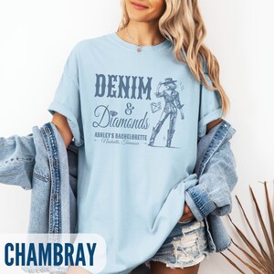 Puede incluir: Una camiseta azul claro con el texto "Denim & Diamonds" y un gr&aacute;fico de una mujer con un sombrero de vaquero y botas. El texto debajo del gr&aacute;fico dice "Ashley's Bachelorette - Nashville, Tennessee". La camiseta est&aacute; hecha de una tela suave y c&oacute;moda y es perfecta para una despedida de soltera.