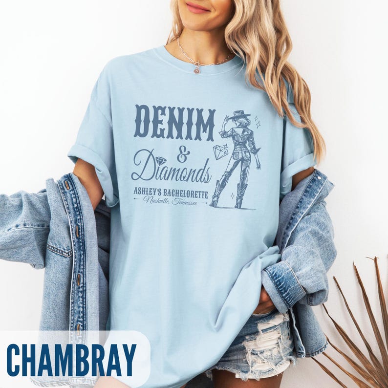 Puede incluir: Una camiseta azul claro con el texto "Denim & Diamonds" y un gr&aacute;fico de una mujer con un sombrero de vaquero y botas. El texto debajo del gr&aacute;fico dice "Ashley's Bachelorette - Nashville, Tennessee". La camiseta est&aacute; hecha de una tela suave y c&oacute;moda y es perfecta para una despedida de soltera.