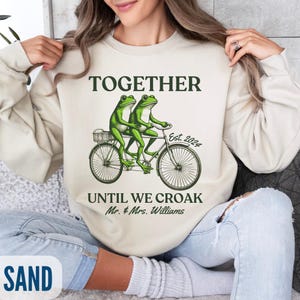 May include: Sand-colored sweatshirt with a whimsical design. Features two green frogs riding a bicycle, with the text "TOGETHER UNTIL WE CROAK" and "Est. 2024". Includes the names "Mr. & Mrs. Williams".