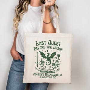 K&ouml;nnte beinhalten: Cremefarbene Tragetasche mit gr&uuml;nem Grafikdesign. Das Design zeigt einen Drachen, ein Schwert, Rosen und den Text "Last Quest Before The Dress" und "Ashley's Bachelorette Charleston, SC".