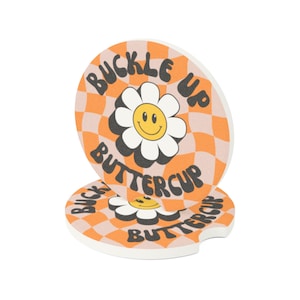 May include: Two round car coasters with a retro design. The coasters have an orange and white checkered pattern with the words "BUCKLE UP BUTTERCUP" in black. A white daisy with a yellow smiley face is in the center.