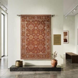 May include: A large, rectangular, rust-colored rug with an intricate floral pattern is displayed on a white wall. The rug is the focal point of a gallery space, complemented by framed artwork, a bench, and decorative items.