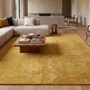 May include: A large, rectangular, mustard-yellow area rug featuring a grid pattern of small, white, cross-shaped tufted accents. The rug has a soft, plush texture and covers a significant portion of the light-coloured wooden floor in the living room.