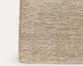 Puede incluir: Primer plano de una alfombra rectangular beige tejida. La alfombra tiene una superficie texturizada con líneas horizontales visibles. Los bordes están cuidadosamente acabados con un borde más oscuro, creando una forma definida. La alfombra está hecha de fibras naturales.