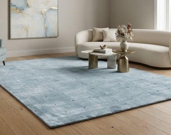 May include: A large, rectangular area rug in a soft, muted blue tone featuring a subtle, grid-like pattern of small, darker dots across its surface. The rug is positioned on a light-colored wooden floor in a room with a sofa and chair.